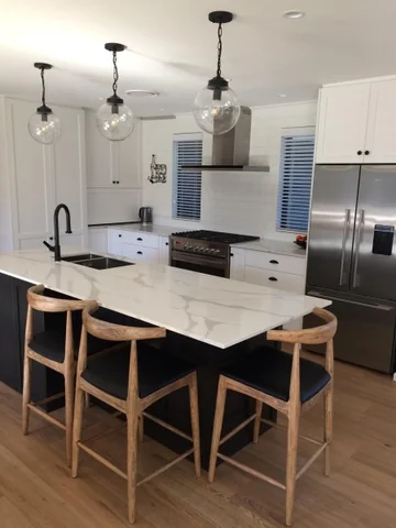 Pale veined island with black cabinetry, timber stools and pendant lighting.